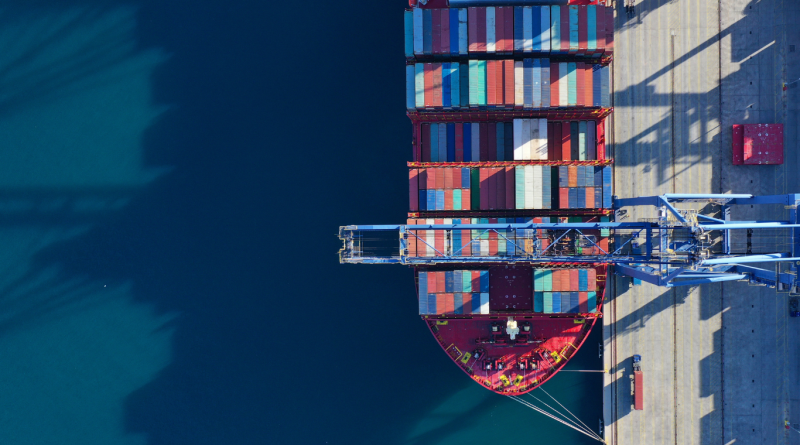 A top-down view of a cargo ship loaded with colorful shipping containers docked at a port, representing supply chain logistics and cost management.