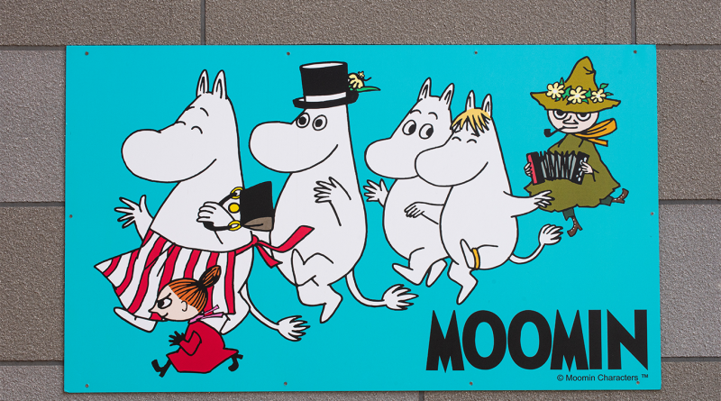 Colorful Moomin characters walking together on a turquoise sign, including Moomintroll, Moominpappa in a top hat, Moominmamma, Little My running in front, and Snufkin playing an accordion.