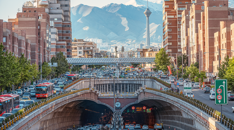 Busy Tehran city street with heavy traffic entering a road tunnel, surrounded by tall buildings, with the Milad Tower and snow-capped mountains visible in the background.