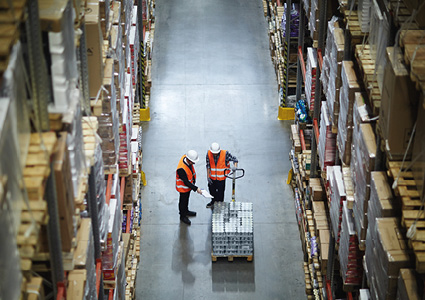 a high-angle, top-down view captures two warehouse workers managing inventory in a central aisle of a large fulfillment center