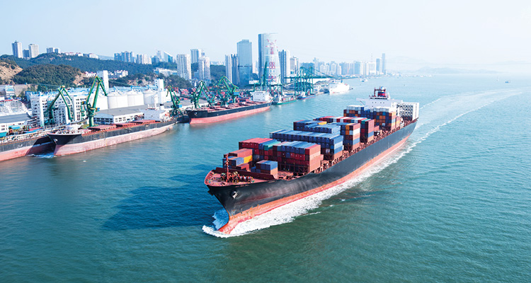 a high-angle aerial view captures a massive container ship cruising through coastal waters away from a bustling deep-water industrial port