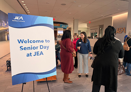 A large white easel sign stands on the left side of the foreground. It displays bold blue letters reading "Welcome to Senior Day at JEA".