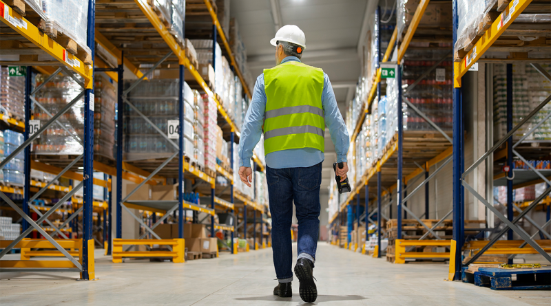 A worker in a safety vest and hard hat walks down the aisles of a warehouse