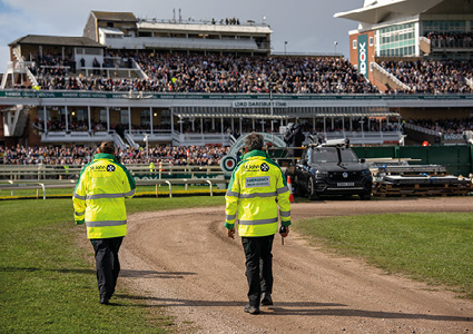 two St John Ambulance personnel in high-visibility jackets walking along a dirt track at a packed horse racing venue