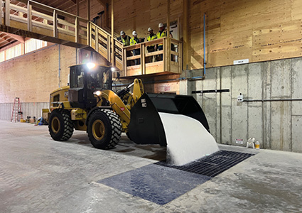 A yellow compact wheel loader is moving bulk material inside an industrial warehouse