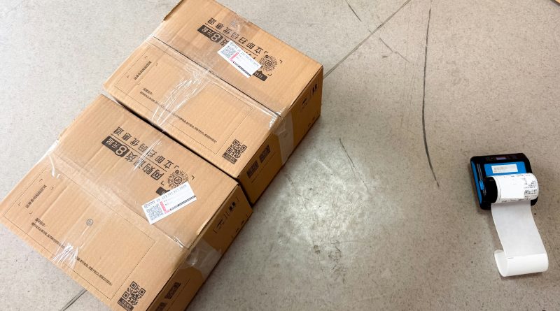 Cardboard shipping boxes with barcode labels beside a portable label printer in a warehouse.